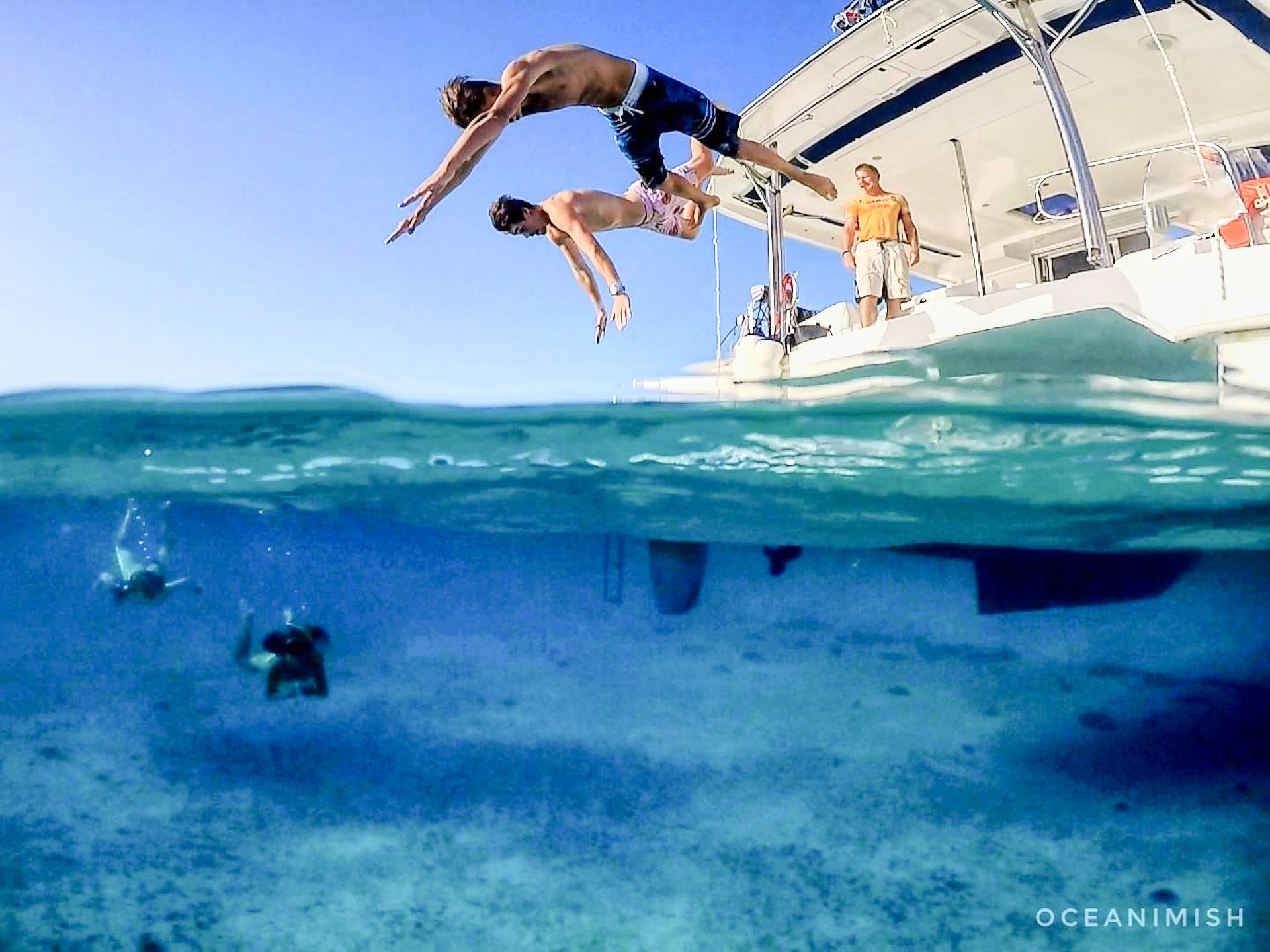 Two teens diving from an Odyssey Expeditions catamaran into clear Caribbean water with snorkelers visible below the surface